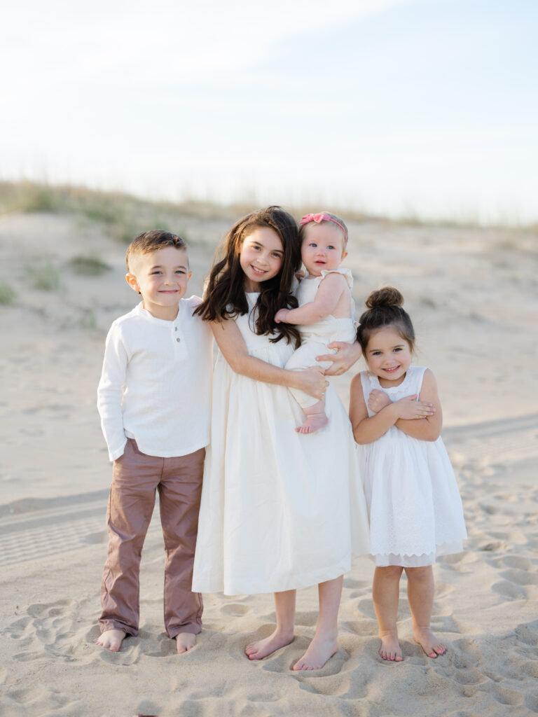 Four kids dressed in white and tan standing on beach by Norfolk family photographer Ashley Brickner 