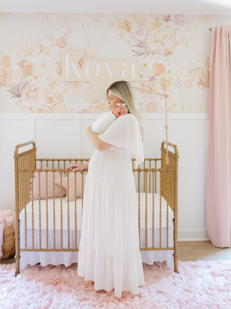 Mother in white dress holding swaddled newborn baby girl in floral pink nursery 
