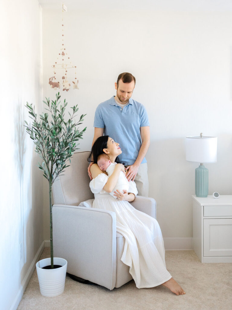 Mother in white dress holding newborn swaddled baby boy while dad looks on  