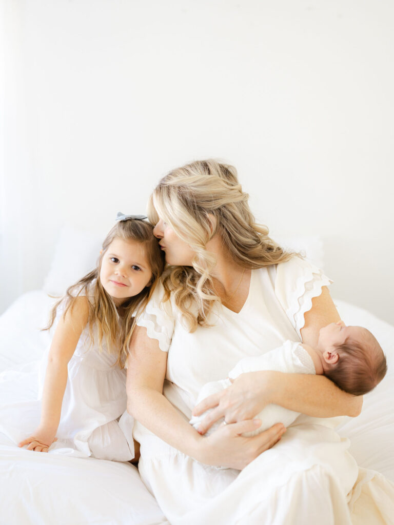 Mom holding newborn baby boy and kissing toddler girl on white bed by Hampton Roads newborn photographer Ashley Brickner 