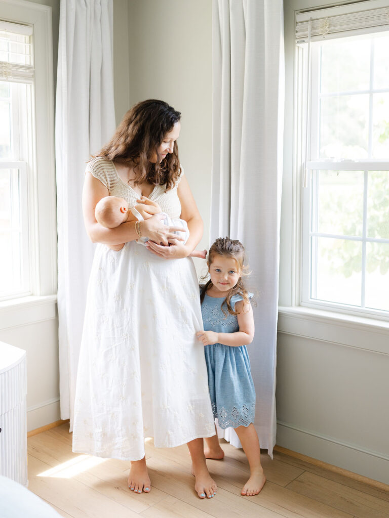 Mother holding baby boy in bedroom with daughter in blue dress looking over during their Virginia Beach newborn session