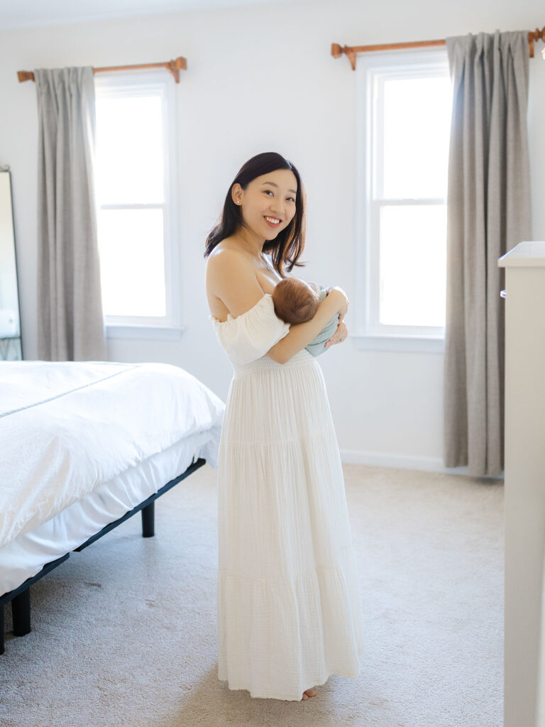 Mother in white dress holding baby boy in her master bedroom during Virginia Beach newborn session