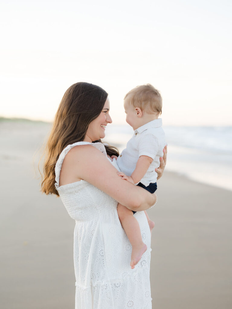 Mom dressed in white holding toddler boy standing on beach for Sandbridge beach photos 