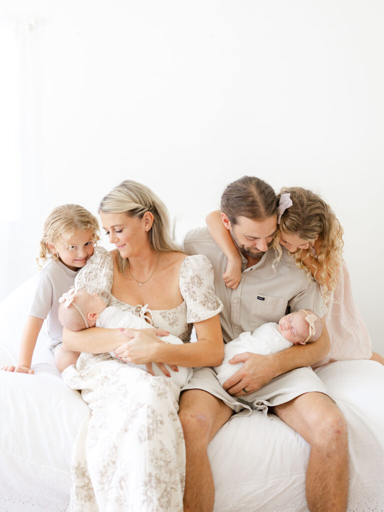 Family of five in neutral clothing sitting on white bed in studio