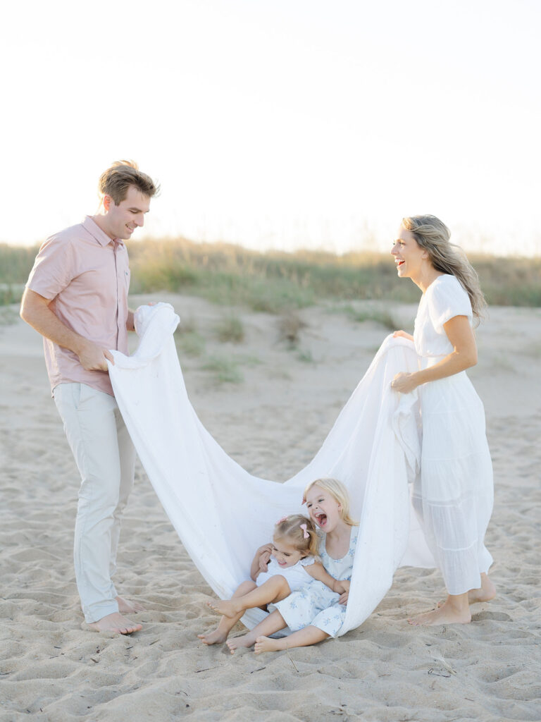 Two parents in neutral clothes holding white blanket with toddler girls laughing in it on the beach by Virginia Beach Family portraits photographer Ashley Brickner