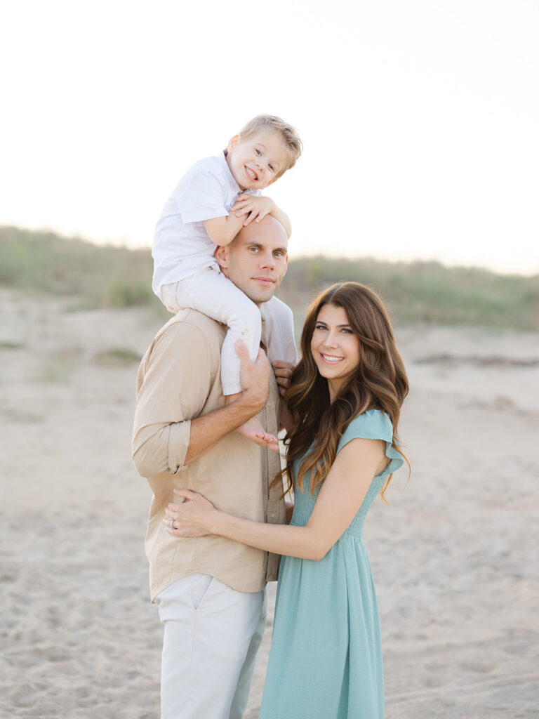 Family of three with toddler son on dad's shoulders standing on the beach by Virginia Beach family portraits photographer Ashley Brickner
