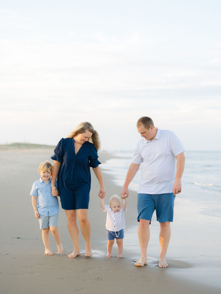 Family of four dressed in blue and white holding hands on the beach by Virginia Beach family portraits photographer Ashley Brickner