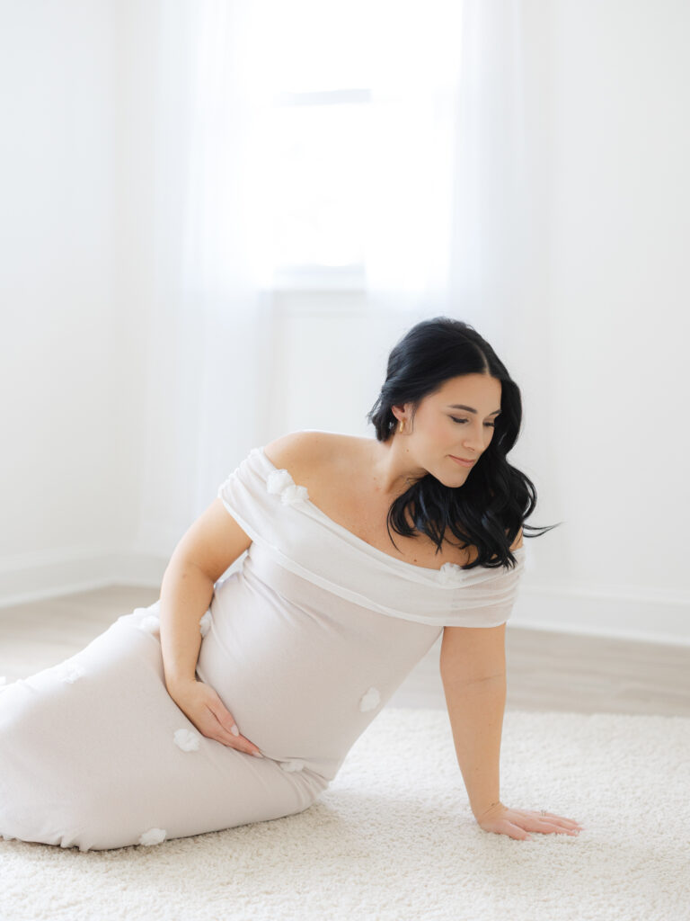Pregnant mom holding belly wearing white dress sitting in studio by Virginia Beach Maternity Portraits Photographer Ashley Brickner 