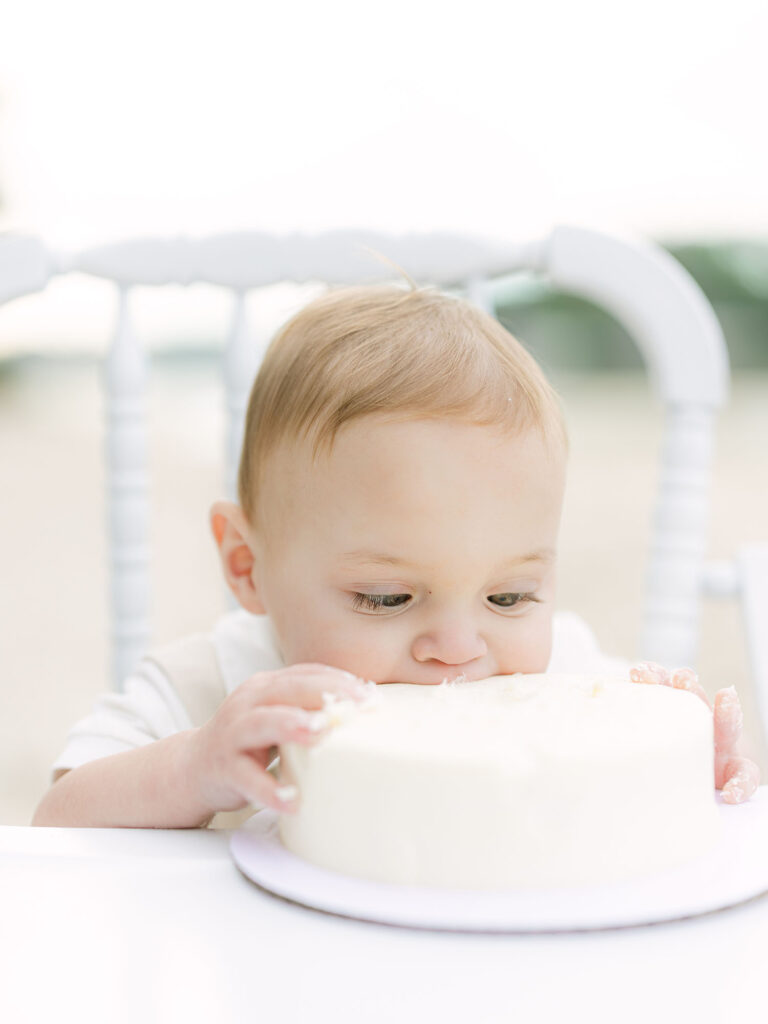Toddler boy biting into white cake in high chair on the beach by photographer who specializes in Hampton Roads cake smash photography