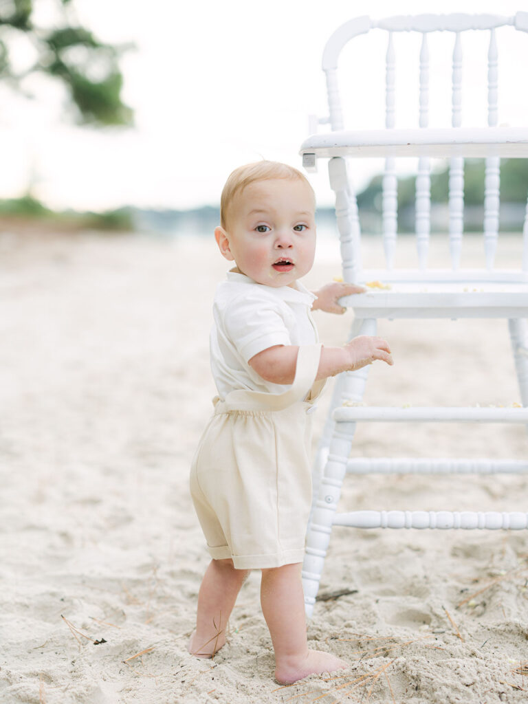 Toddler boy standing on beach leaning on high chair by photographer who specializes in Hampton Roads cake smash photography