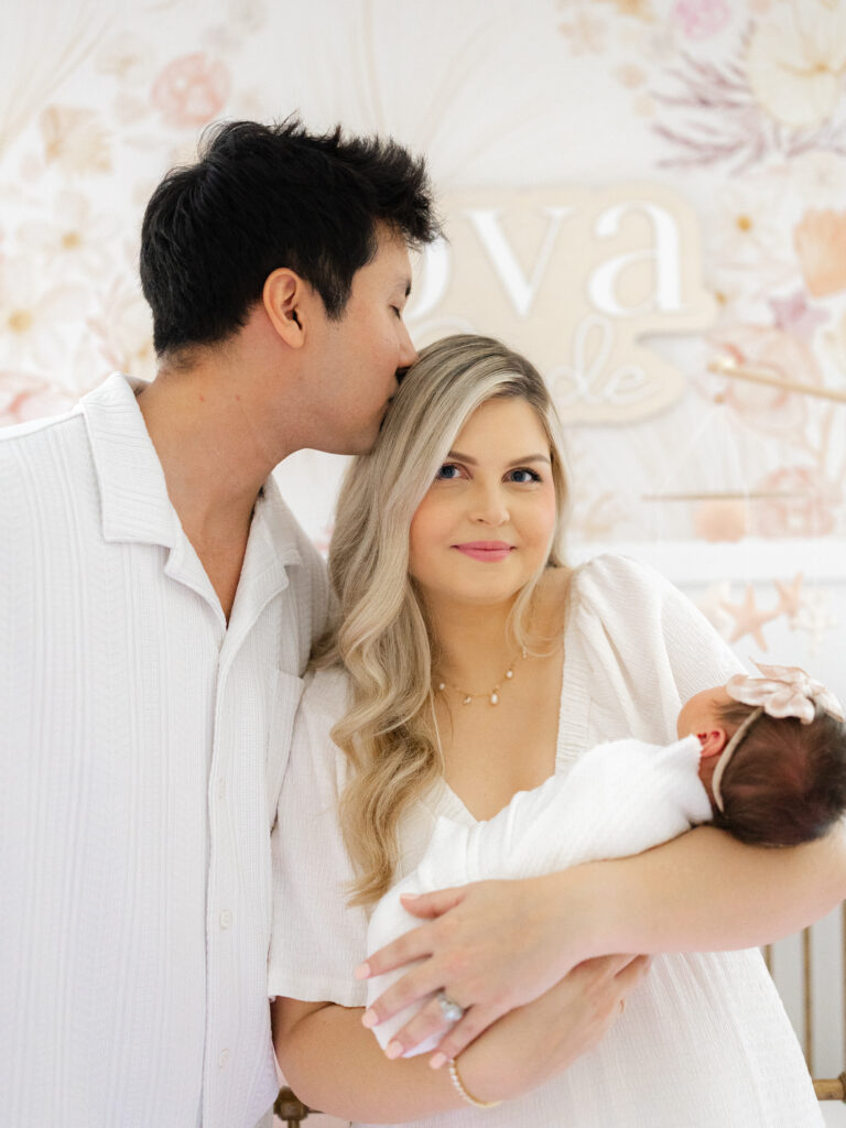 Dad kissing mom holding newborn baby girl on the head in floral pink nursery for an in-home lifestyle newborn session by Ashley Brickner 