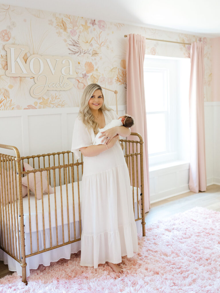 New mom standing in pink and floral nursery holding newborn baby girl for an in-home lifestyle newborn session by Ashley Brickner 