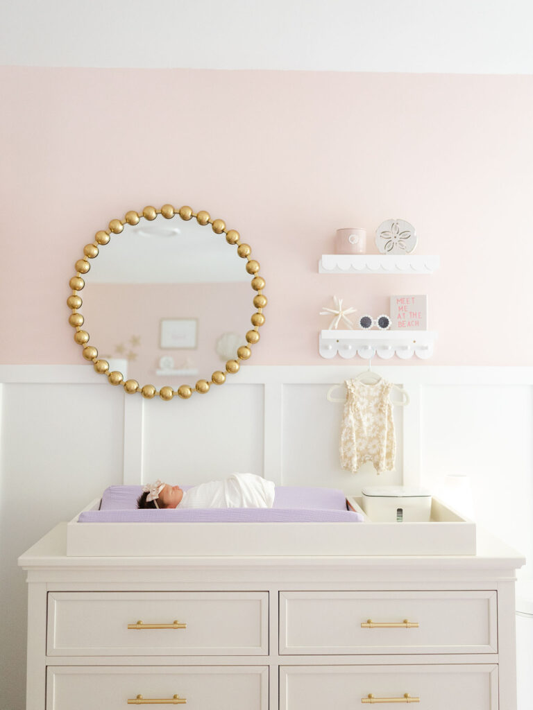 Baby girl laying on white dresser with gold pulls and gold mirror above photographed for an in-home lifestyle newborn session by Ashley Brickner 