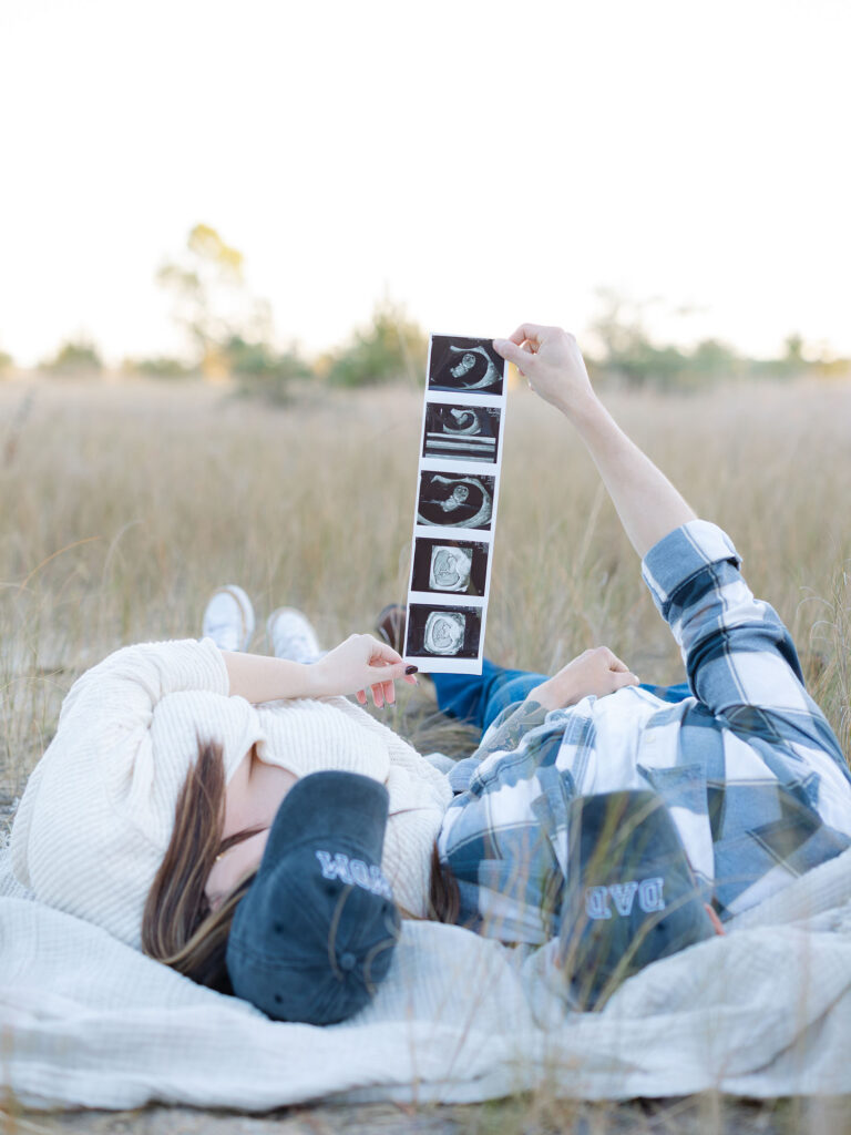 Husband and wife laying in Pleasure House Point field holding ultrasound photo 