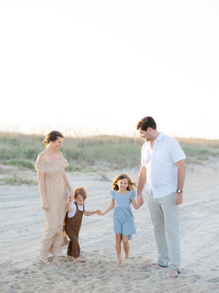 Family of four dressed in tan and blue holding hands and standing on the beach taken by Virginia family photographer Ashley Brickner 