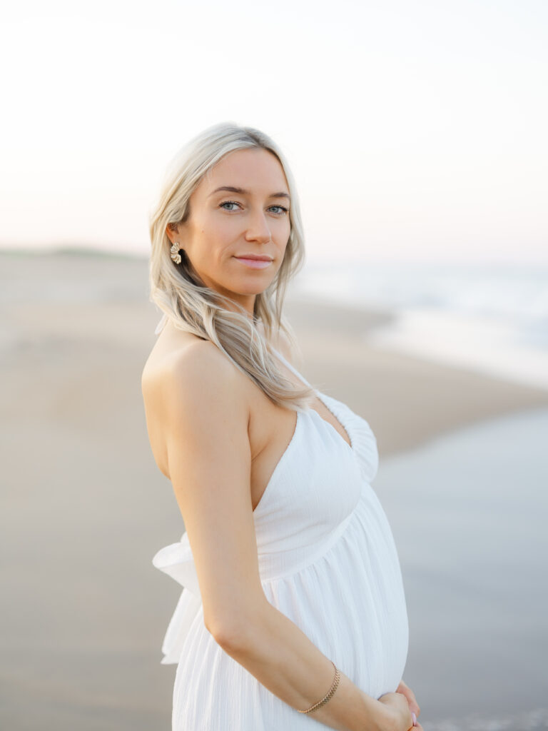Pregnant mom holding belly in white dress standing on beach by Virginia Maternity Photographer Ashley Brickner 