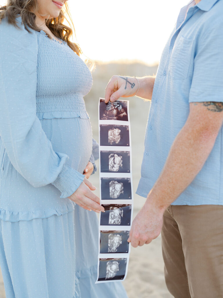 Mom and dad dressed in blue on beach holding ultrasound photo 