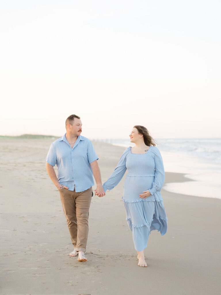 Husband and wife dressed in blue and walking while holding hands on the beach by Virginia Maternity Photographer 