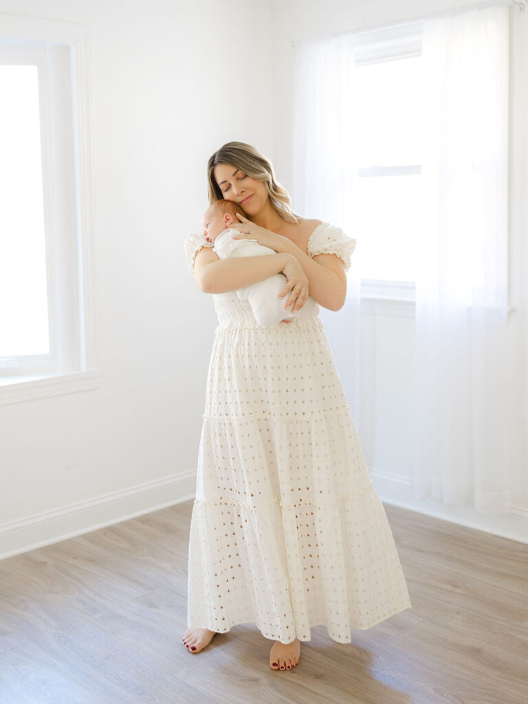 Mom in white maxi dress rocking and holding swaddled baby boy in studio 