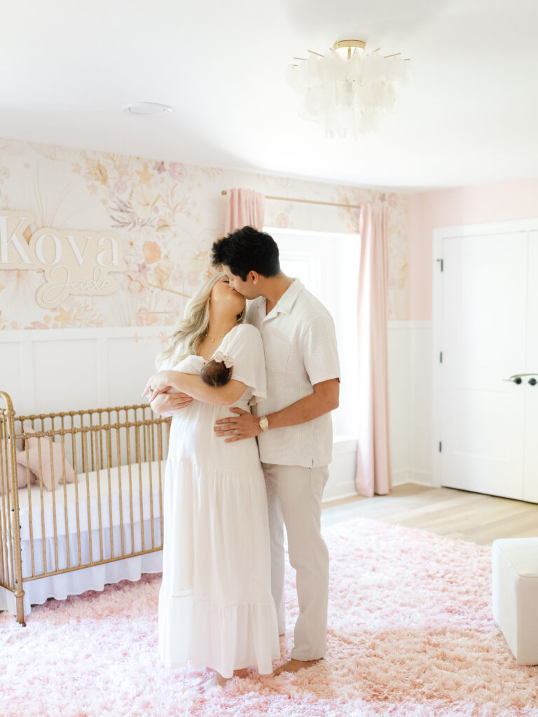 Mom and dad kissing and standing in nursery while holding newborn baby girl by Virginia newborn photographer Ashley Brickner 