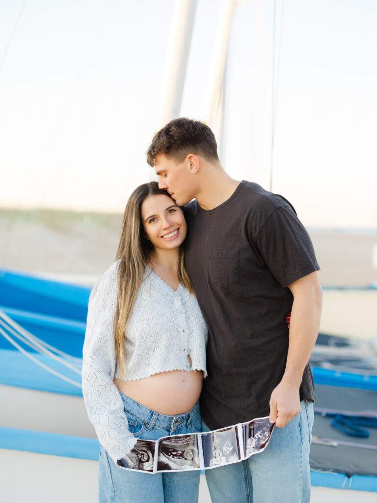 Pregnant mom and husband holding ultrasound photos on beach in front of blue boat by Ashley Brickner