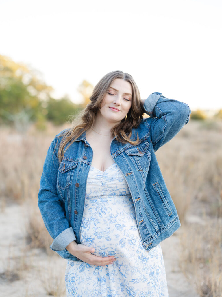 Pregnant mom in blue floral dress and denim jacket holding belly and standing in a field by Ashley Brickner
