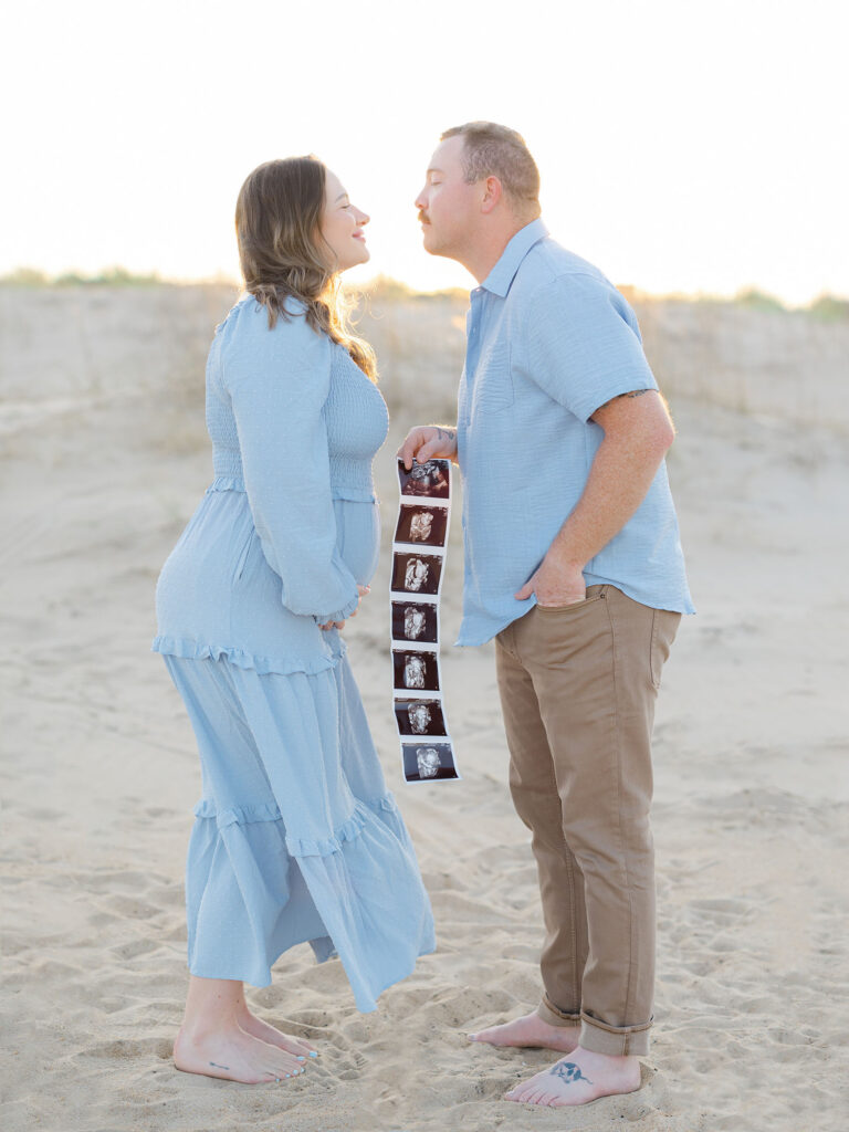 Pregnant mom and dad in blue and tan holding ultrasound photos standing on the beach by Ashley Brickner