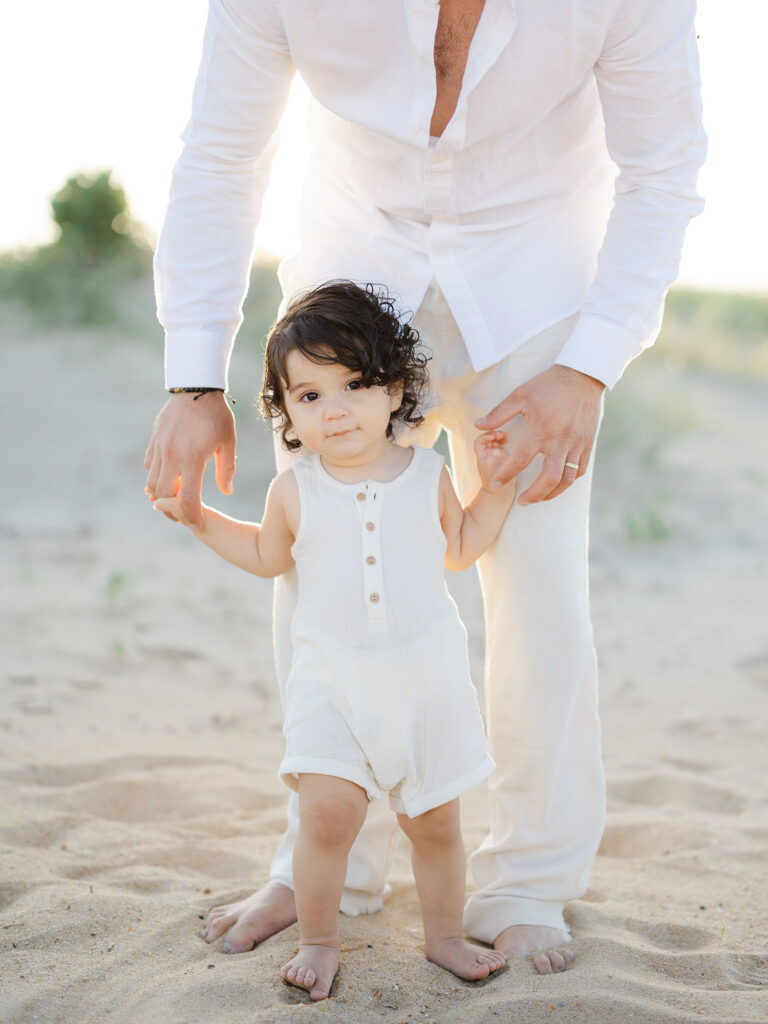 Dad standing on beach holding hands with toddler boy on beach 