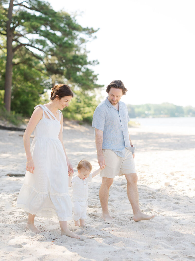 Mom dad and toddler boy in neutral clothes walking on beach 