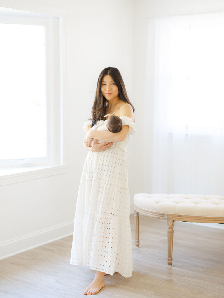 Mom in white dress standing in studio holding swaddled newborn baby girl during milestone session photography by Virginia Beach photographer Ashley Brickner 