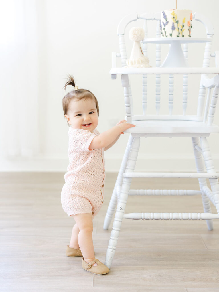 Baby girl standing and holding high chair with cake on it in Virginia Beach studio 