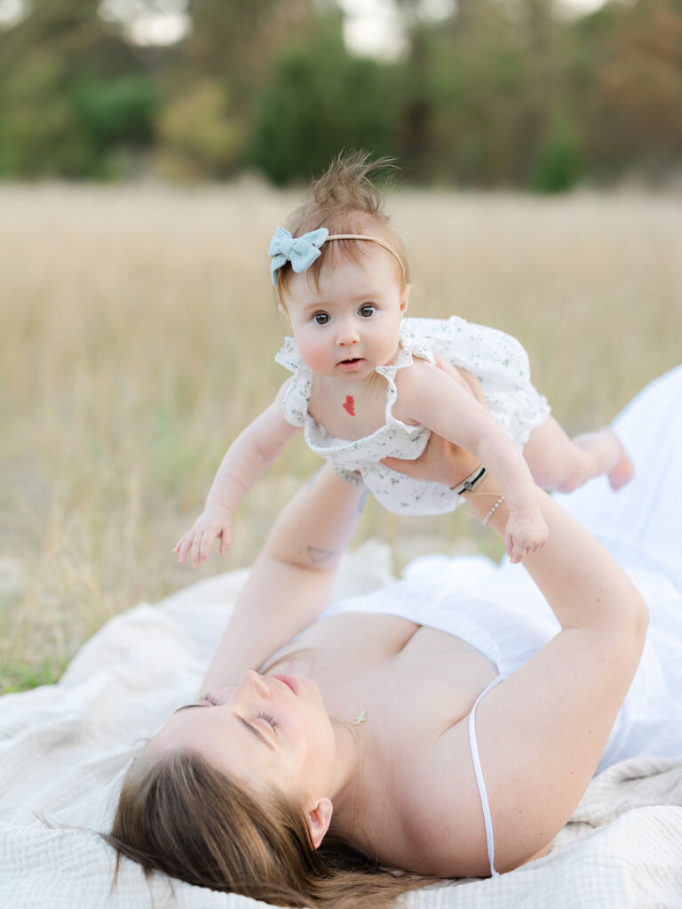 Mom in white dress holding baby girl up in field during milestone session photography by Virginia Beach photographer Ashley Brickner 