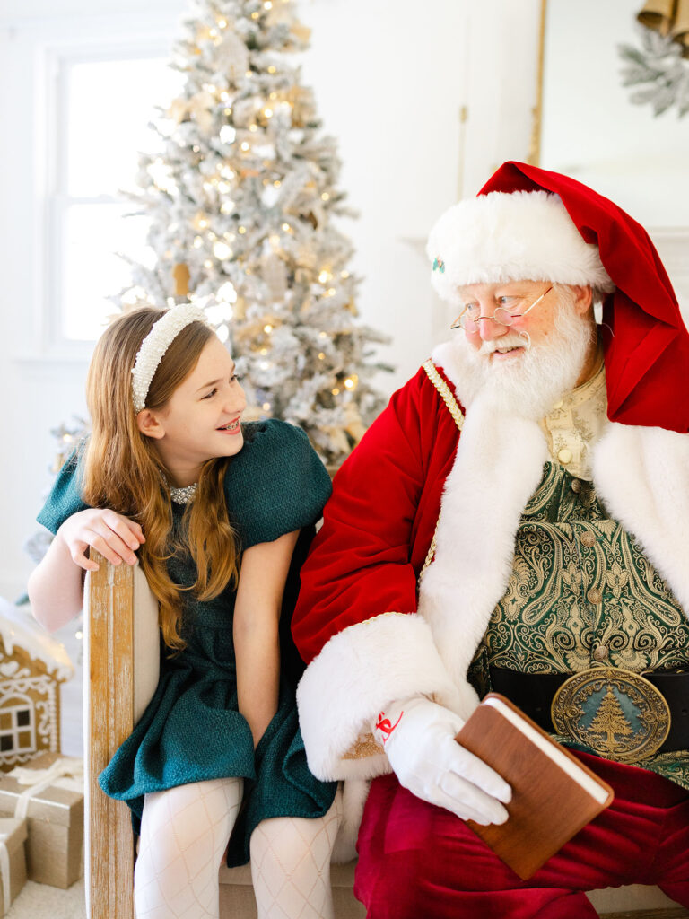 Santa and girl in green sitting in natural lit studio with holiday setup 