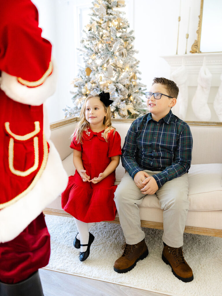 girl and boy dressed in christmas colors and looking up at santa with holiday setup behind them 